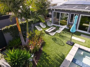 A photo of a beautiful backyard area with six chairs, an umbrella, artificial turf grass, afire pit, and a pool. 