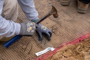 image of hands with work gloves holding a hammer and nail over gopher wire. 